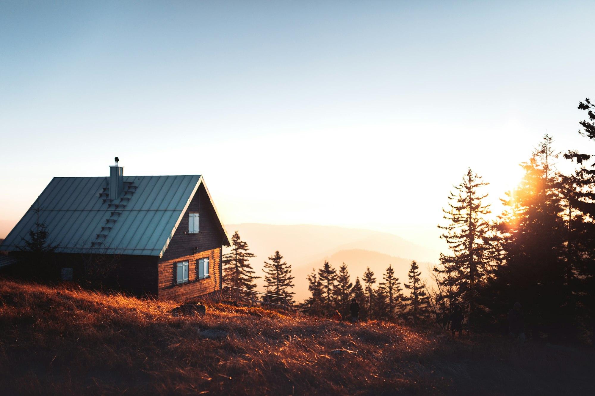A wooden house on a hillside at sunset, with pine trees behind it